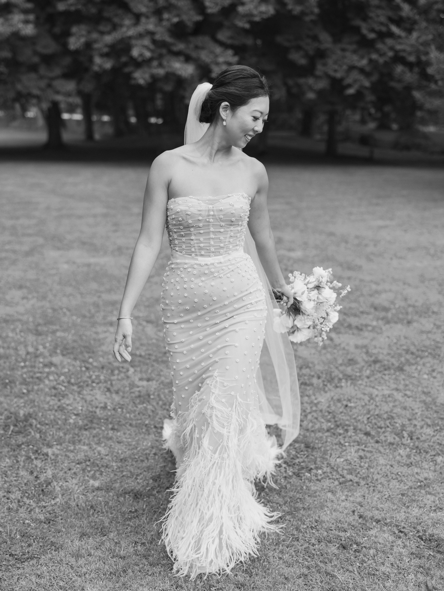 A bride in a strapless, embellished gown with a feathered skirt holds a bouquet, walking in a park, captured in black and white at Chateau Allure du Lac Loire VAlley