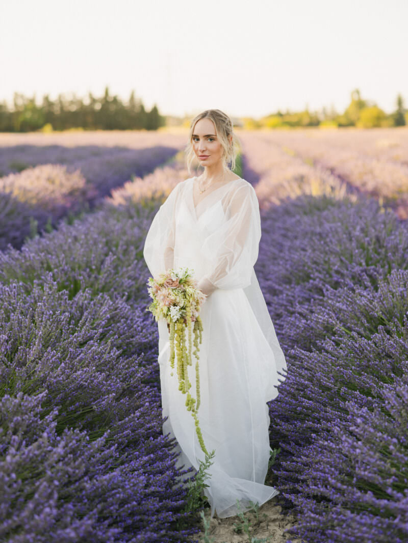 A bride in a translucent white gown stands amidst a vibrant lavender field in Provence, holding a bouquet in the surroundings of Bastide Saint Antoine.
